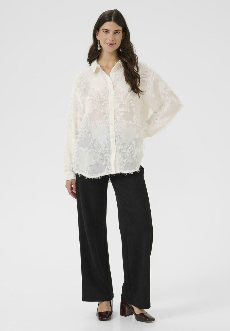 Prozorna bela bluza z reliefnim cvetličnim vzorcem, gumbe na sprednji strani in dolgimi rokavi, v kombinaciji s črnimi širokimi hlačami in bordo petami.