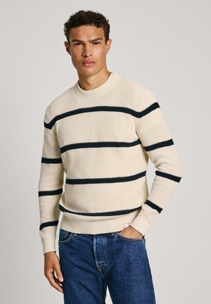 Maglione a maglia crema con strisce orizzontali blu scuro, scollatura rotonda, polsini e orlo a coste. Abbinato a jeans di denim blu.