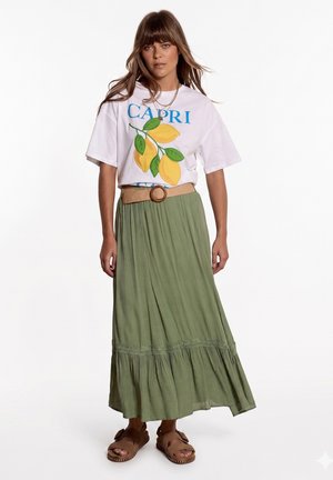 Frau trägt weißes T-Shirt mit Zitronenprint, olivgrünen langen Rock mit Gürtel und braune Sandalen, steht vor einfachem weißem Hintergrund.
