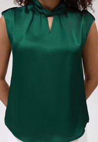 Blusa satiné verde con un design a occhiello sul davanti, maniche corte e un dettaglio a fiocco sul collo. Tessuto liscio, leggermente oversize.