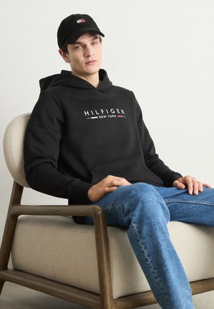 Sudadera negra con el logo "HILFIGER NEW YORK" en blanco, rojo y azul. Llevada con jeans de color azul claro, sentado en una silla de color claro. Gorro negro.