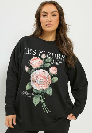 Femme portant un sweat-shirt noir avec un bouquet de roses roses et un texte blanc indiquant "LES FLEURS STUDIO PARIS" à l’avant.