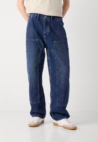 Jean en denim bleu avec une coupe décontractée, comportant de grandes poches avant et des coutures contrastées. Portés avec des baskets blanches sur un fond clair.