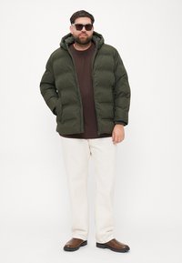 Puffer jacket in dark green with a hood, featuring a zip closure, worn over a brown shirt and light cream pants, paired with brown shoes.