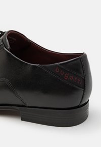 Zapato de vestir de cuero negro con un acabado liso, que presenta un bordado intrincado y un logo rojo "bugatti" en el lateral. Diseño de tacón bajo.