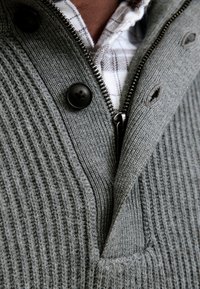 Grey ribbed pullover with a zipper, featuring two black buttons and a collar. Underneath, a white checkered shirt is partially visible.