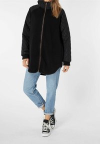 Giacca in pile nera con maniche in nylon trapuntato nero, colletto alto e zip frontale. Indossata con jeans azzurri arrotolati e scarpe da ginnastica nere.