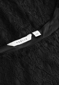 Fekete csipkebőrből készült ruha sima selyemnyakkal. Bonyolult csipke mintákkal rendelkezik. A címke azt írja, hogy "NOELLA", valamint a méret és az eredet országának részleteit tartalmazza.