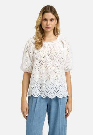 Woman wearing white eyelet puff-sleeve blouse with scalloped hem and light blue high-waisted wide-leg jeans, standing against plain background.