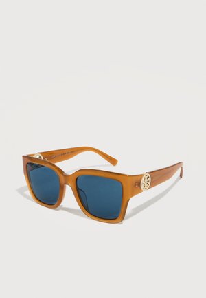 Lunettes de soleil carrées translucides marron avec des verres bleu foncé et un logo décoratif doré sur les branches, présentées sur un fond blanc.