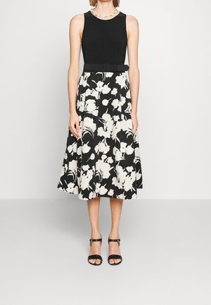 Kobieta ubrana w czarny top bez rękawów i czarną spódnicę z wysokim stanem w białe kwiatowe wzory, czarny pasek, czarne szpilki z paskiem wokół kostki oraz perłowy naszyjnik.