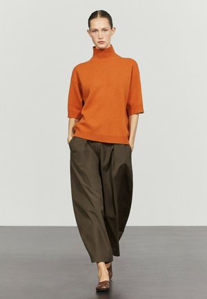 Donna in piedi che indossa un maglione arancione a collo alto e maniche corte, pantaloni larghi marrone scuro e scarpe piatte marroni su uno sfondo uniforme.