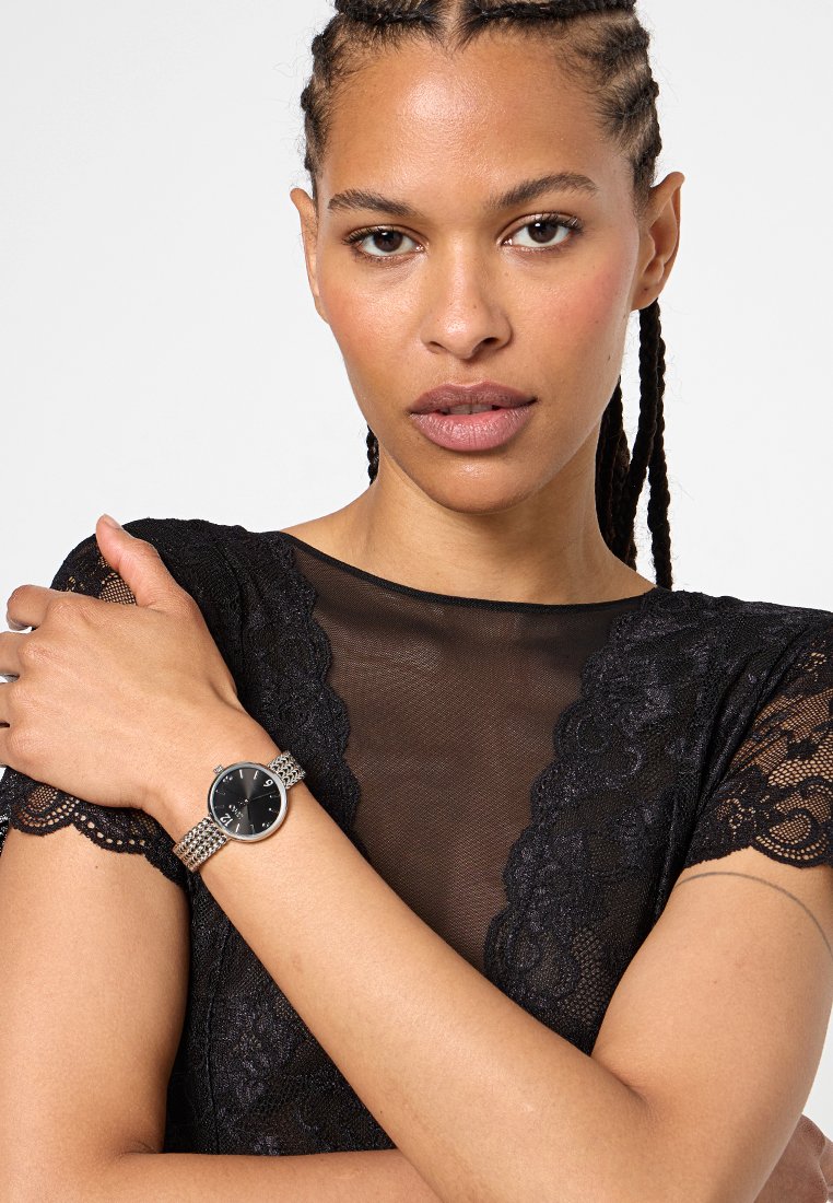 Srebrny zegarek na nadgarstek z okrągłą czarną tarczą, siateczkowym paskiem i białymi oznaczeniami godzin. Noszony na nagim ramieniu, w połączeniu z czarną koronkową bluzką.