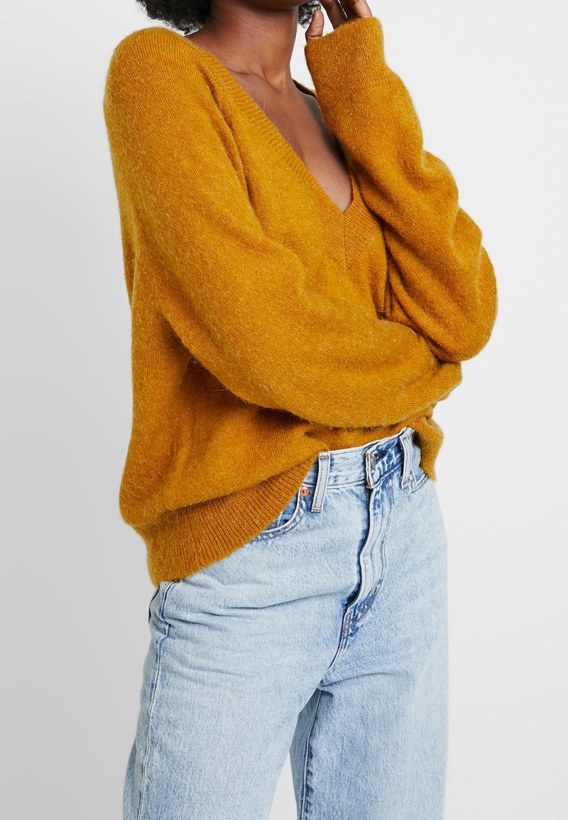 Pull oversize jaune moutarde à col en V, confectionné dans une matière douce. Associé à un jean taille haute bleu clair.