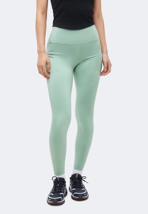 Άτομο που φοράει ψηλόμεσα τιρκουάζ κολάν, μαύρο αμάνικο τοπ, λευκές κάλτσες και μαύρα αθλητικά παπούτσια, στέκεται σε μονόχρωμο φόντο.