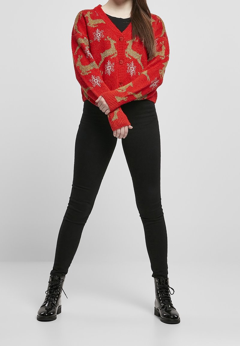 Röd stickad tröja med guldrenar och snöflingemönster, med V-hals, knäppning och förlängda ärmmuddar. Kombineras med svarta leggings och stövlar.