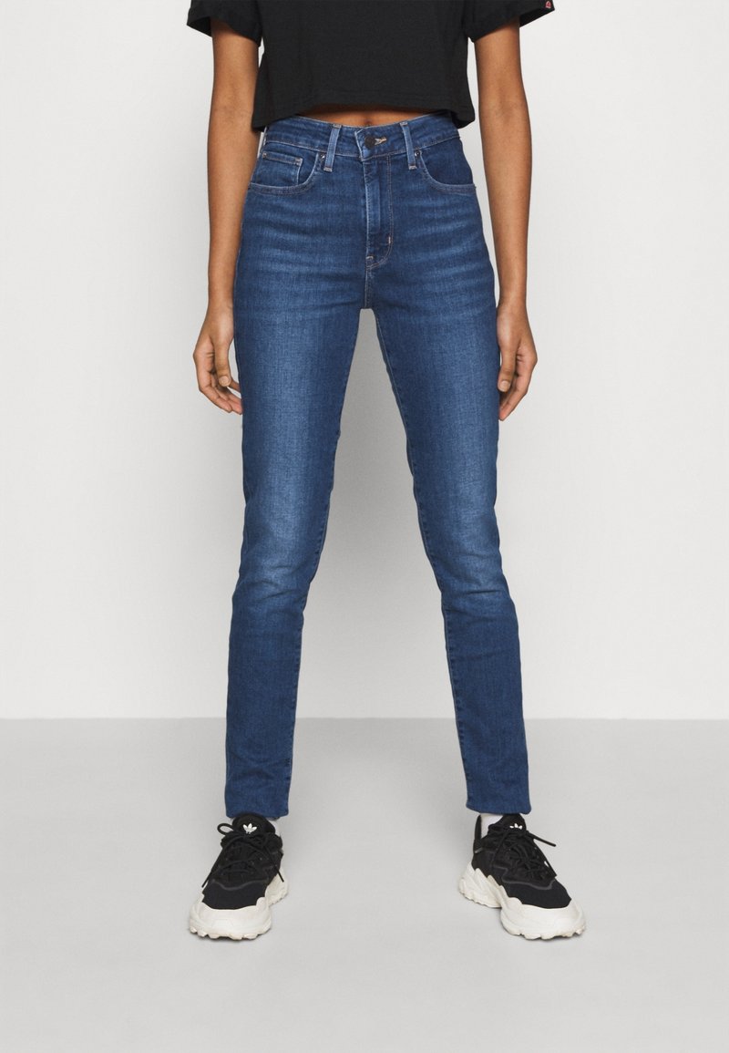 Hochtaillierte, schmal geschnittene, blaue Jeans mit einer glatten Denim-Oberfläche und einem Fünf-Taschen-Design, kombiniert mit schwarzen Sneakers und einem cropped Top.
