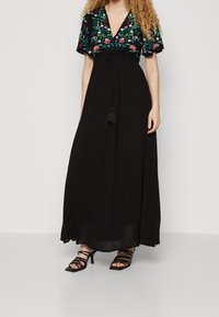 Schwarzes Maxikleid mit floralem Stickerei auf einem kurzärmeligen Oberteil, mit einem Bindeband in der Taille und einem fließenden Rock. Sichtbare schwarze Riemchen-Absätze.