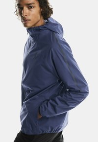 Giacca a vento con cappuccio da uomo in blu scuro, caratterizzata da una texture liscia, chiusura con zip, con dettagli riflettenti lungo le maniche e un design aderente.
