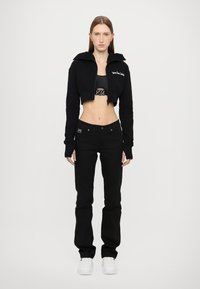 Melna īsā kapuce ar augstu apkakli, saskaņota ar melnām pieguļošām džinsu biksēm un baltām sporta kurpēm. Iezīmēta ar jostu ar metāla akcentiem.