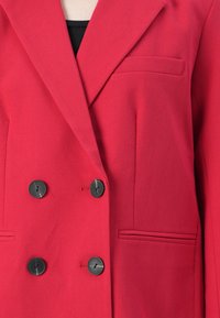 Červený dvouřadový blazer se čtyřmi černými knoflíky a třemi kapsami, nošený přes černý top.