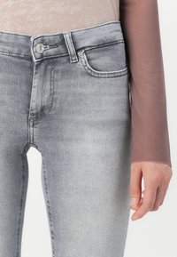 Detailní záběr na šedé vybledlé džíny s knoflíkem a zipem, přední kapsou a pravou rukou osoby odpočívající vedle stehna.