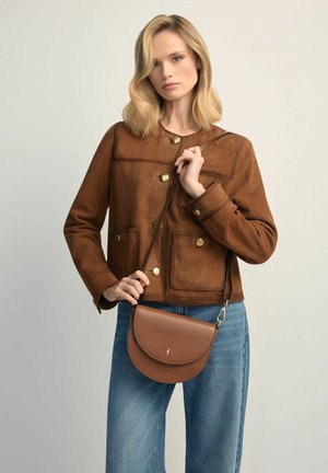 Kobieta ubrana w brązową zamszową kurtkę zapinaną na guziki i niebieskie dżinsy, trzymająca brązową skórzaną torebkę na pasku.