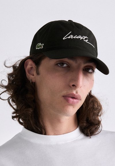 Jeune personne avec de longs cheveux bouclés portant une casquette Lacoste noire et une chemise blanche, faisant face à l'avant avec une expression neutre devant un fond uni.