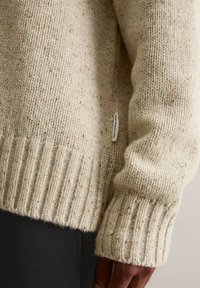 Sweater de malha em bege claro com textura salpicada. Apresenta uma bainha e punhos canelados, com uma pequena etiqueta de logotipo ao lado. Material macio e espesso.