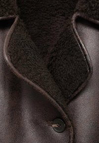 Esterno in pelle marrone con un morbido colletto in pile marrone testurizzato. Presenta una chiusura con bottone nero e finiture lisce lungo i bordi.