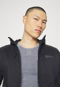 Black hooded jacket with zippered pockets, featuring a textured gray shirt underneath. The jacket has a logo on the left chest.