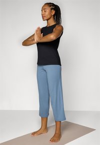 Svart ermeløs topp med skuldersnitt, kombinert med lyseblå cropped bukser. Emnet står på en beige yogamatte, med bare føtter.