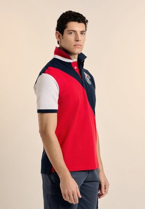 Hombre con cabello corto, rizado y oscuro, vestido con una camiseta tipo polo de rugby en rojo, azul marino y blanco, de pie y mirando ligeramente hacia un lado.