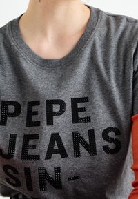 Camiseta de algodón gris con texto negro "PEPE JEANS SIN-" adornada con rhinestones. Cuenta con un acento naranja en la manga. Cuello redondo.