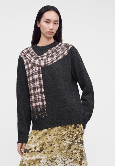 Femme aux longs cheveux noirs portant un pull gris foncé avec un motif d’écharpe à carreaux roses et une jupe à motif d’écailles dorées, brunes et blanches.