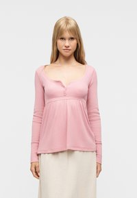 Νεαρή γυναίκα με μακριά ξανθά μαλλιά, που φοράει ροζ μπλούζα με μακριά μανίκια και μπεζ φούστα, κοιτάζοντας προς τα εμπρός μπροστά σε ένα απλό φωτεινό φόντο.