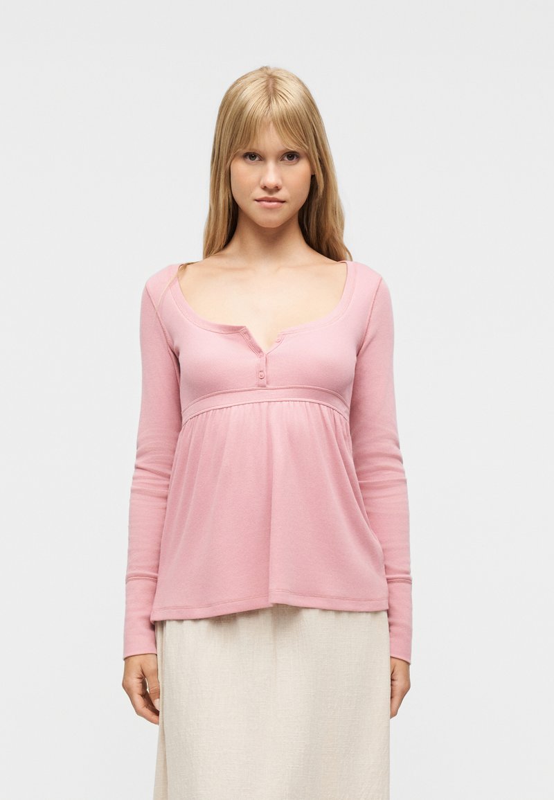 Νεαρή γυναίκα με μακριά ξανθά μαλλιά, που φοράει ροζ μπλούζα με μακριά μανίκια και μπεζ φούστα, κοιτάζοντας προς τα εμπρός μπροστά σε ένα απλό φωτεινό φόντο.