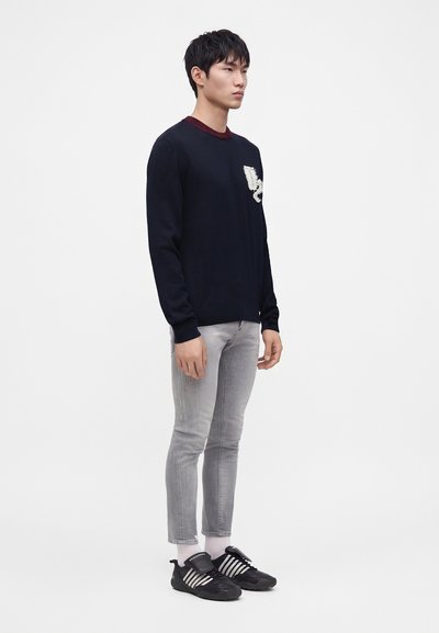 Jeune homme debout portant un pull noir avec un motif blanc, un jean skinny gris clair, des chaussettes blanches et des baskets noires à rayures blanches.