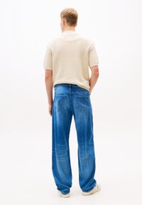 Beige textured short-sleeve shirt paired with relaxed-fit blue denim jeans featuring back pockets and slightly faded areas. Neutral background.