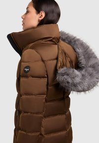 Rjava puhovka z visokim ovratnikom, prešito teksturo, sivim umetnim krznenim robom na kapuci in črno logotipno značko na rokavu.