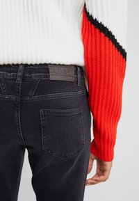 Persona que lleva puesto unos pantalones de mezclilla negros con un bolsillo trasero y un suéter blanco con mangas de rayas rojas y negras, con la mano relajada a su lado.