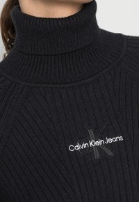 Pull à col roulé noir en matière côtelée, avec un logo discret en gris foncé et blanc : "Calvin Klein Jeans".