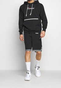 Sudadera negra con capucha y estampado de "Baloncesto", bolsillo con cremallera; combinada con pantalones cortos negros con ribete blanco y logo. Calcetines y zapatillas deportivas blancas.