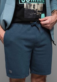 Pantalones cortos de algodón azul con cintura elástica y cordón de ajuste, con bolsillos laterales y un pequeño parche de logo en el dobladillo.
