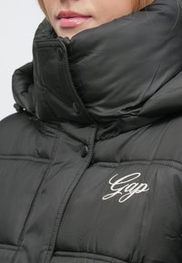 Sort dunjakke med høj krave, med trykknapper og et hvidt broderet "Gap"-logo på brystet. Glat, skinnende tekstur.