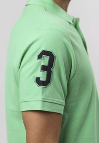 Roheline polo-särk, mille vasakul varrukal on tikitud must number 3. Kangas on tekstureeritud puuvill, klassikalise krae ja lühikeste varrukatega.