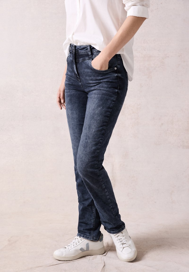 Jeans slim fit in denim scuro con una texture liscia, design a cinque tasche e passanti per cintura, abbinati a sneakers bianche con accenti grigi.
