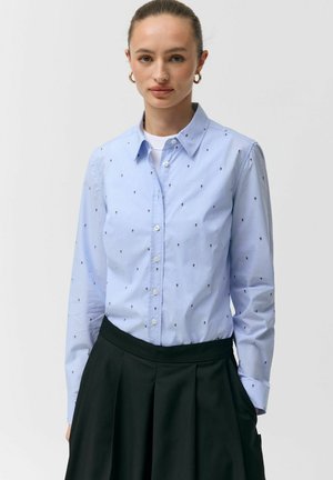 Kobieta ubrana w jasnoniebieską koszulę na guziki z małymi ciemnymi wzorami oraz czarną plisowaną spódnicę, stojąca na jednolitym tle.