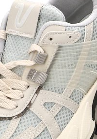 Primer plano de una zapatilla deportiva gris claro con tela de malla, cordones gruesos color crema, superposiciones sintéticas y una correa lateral de plástico transparente.