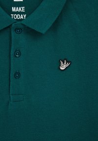 Zöldes-kék póló, textúrált anyagból, három gombbal, a bal mellen fehér és fekete hímzett madárlogóval.